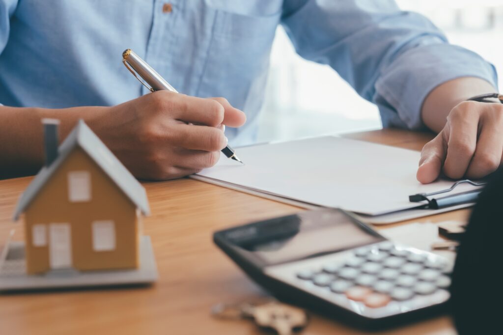 Person signing documents with house model and calculator nearby, representing dental practice sale planning and broker-assisted transitions through Legacy Practice Transitions.